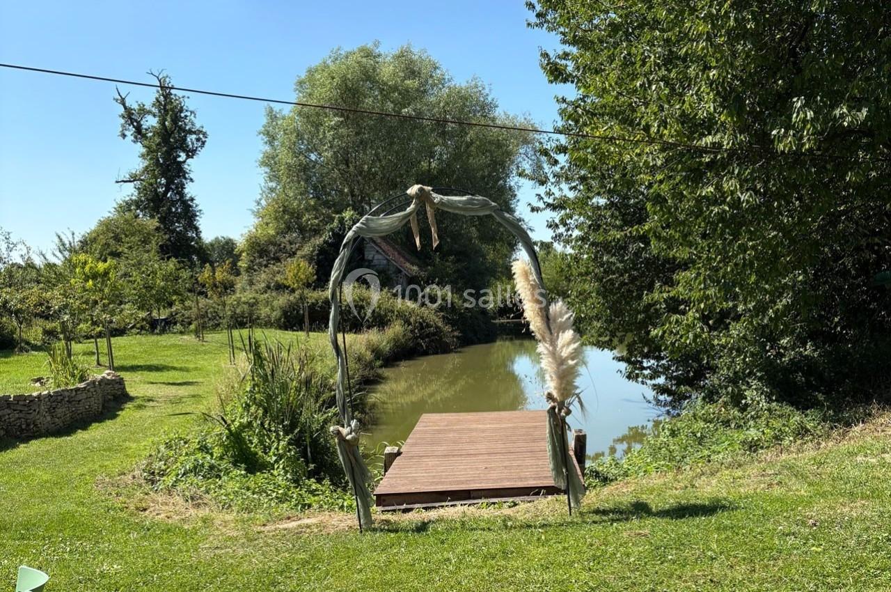 Arche décorative en bois et tissu près d'un ponton au bord d'un étang entouré de verdure.