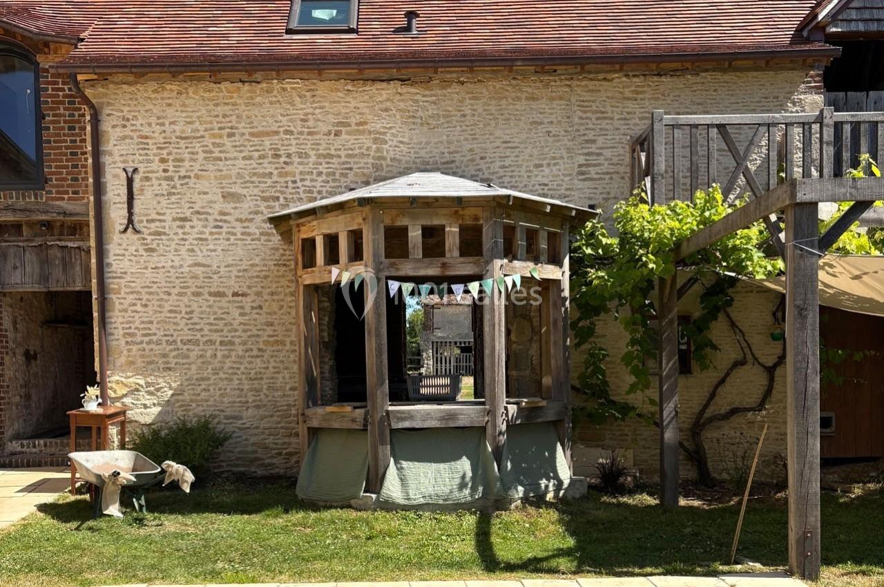 Abri en bois rustique avec guirlande colorée, adossé à une maison en pierre avec toit en tuiles rouges.