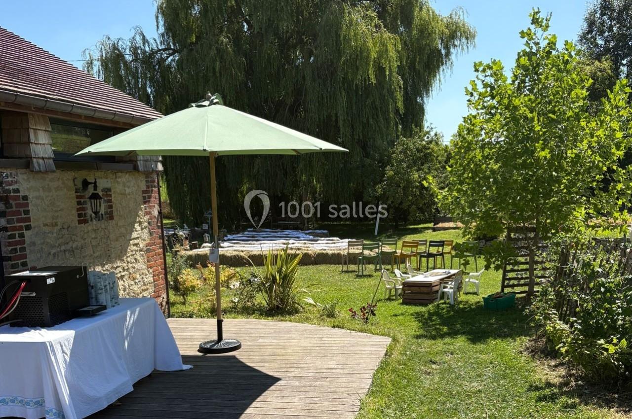 Jardin verdoyant avec terrasse en bois, parasol vert, table dressée et chaises disposées sous un grand arbre.