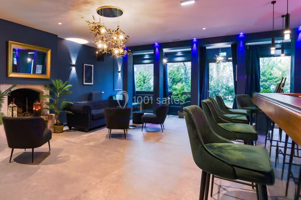 Salle moderne avec fauteuils verts, bar en bois, cheminée allumée et grandes fenêtres donnant sur un jardin.