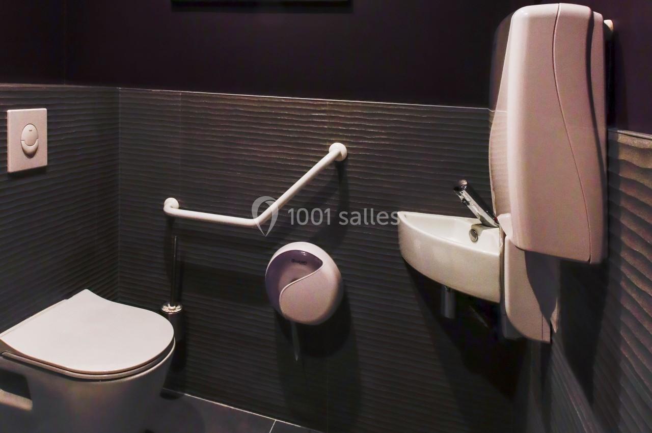 Toilettes accessibles avec barre d'appui, lavabo mural et distributeur de savon dans un espace sombre.