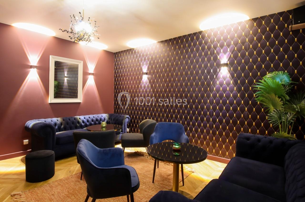 Salon élégant avec murs violets à motifs dorés, canapés en velours bleu, tables noires et éclairage tamisé.