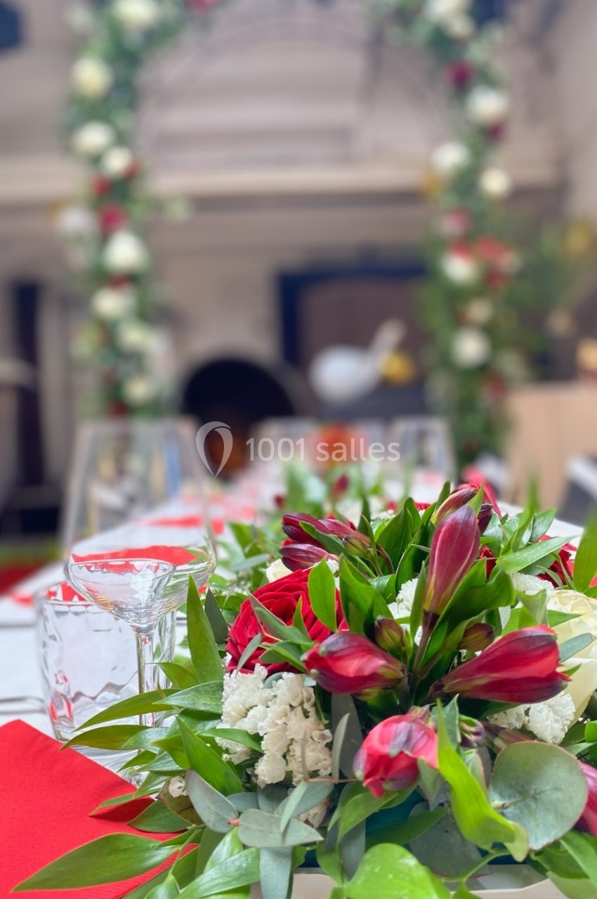 Centre de table floral avec des fleurs rouges et blanches, entouré de verres et nappes rouges, décor d'arche en arrière-plan.