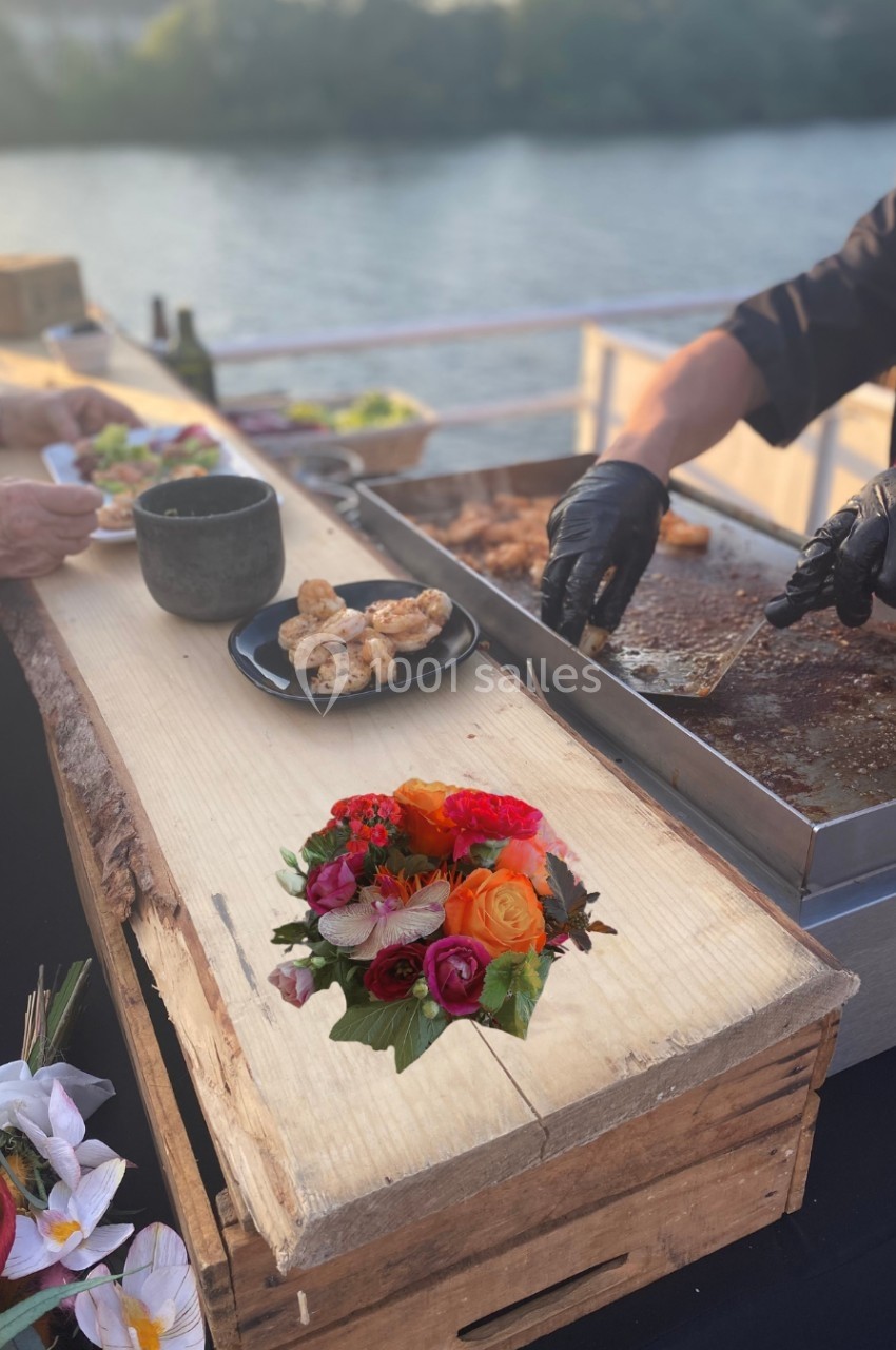 Planche en bois avec fleurs en premier plan, stand de cuisine en extérieur avec grillades et vue sur l'eau en arrière-plan.