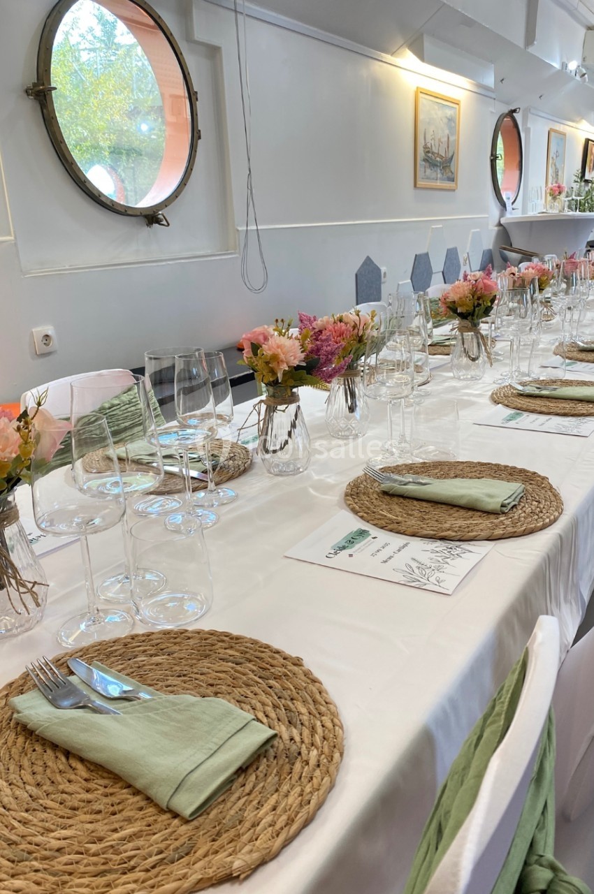 Table dressée avec nappes blanches, sets en osier, serviettes vertes, verres et bouquets de fleurs colorées dans une salle…