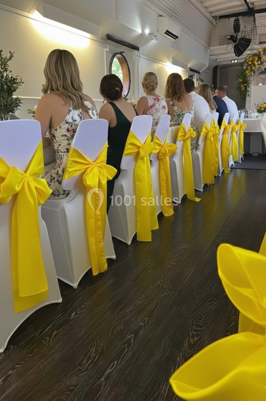 Chaises blanches décorées de nœuds jaunes alignées autour d'une table lors d'un événement festif en intérieur.