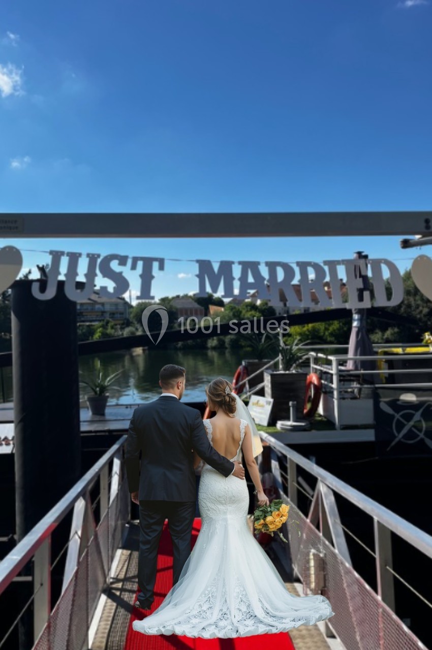 Un couple en tenue de mariage marche sur un ponton décoré avec une banderole ’Just Married’.