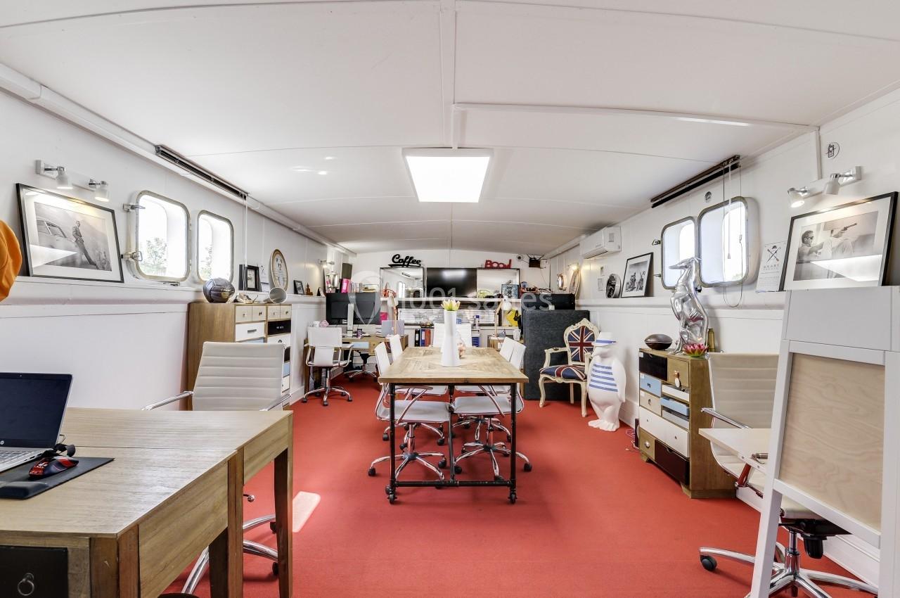 Intérieur lumineux d'un bureau aménagé dans une péniche, avec mobilier moderne, murs blancs et sol rouge.