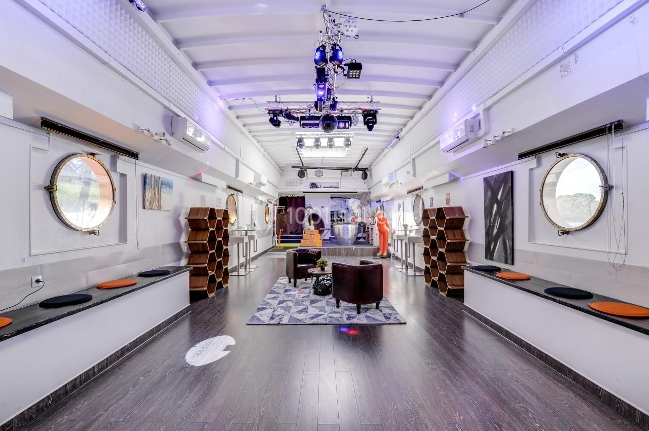 Salle lumineuse avec parquet sombre, fauteuils, étagères hexagonales, hublots et éclairage suspendu moderne.