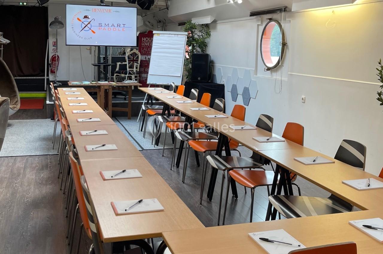 Salle de réunion aménagée avec des tables en U, des chaises, des blocs-notes, un écran de présentation et un paperboard.