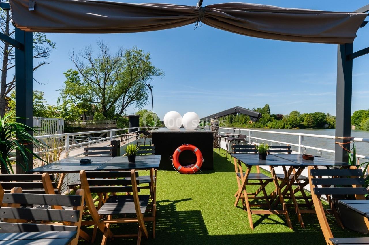 Terrasse extérieure avec tables, chaises en bois, pelouse artificielle, bouée rouge et vue sur une rivière bordée d'arbres.