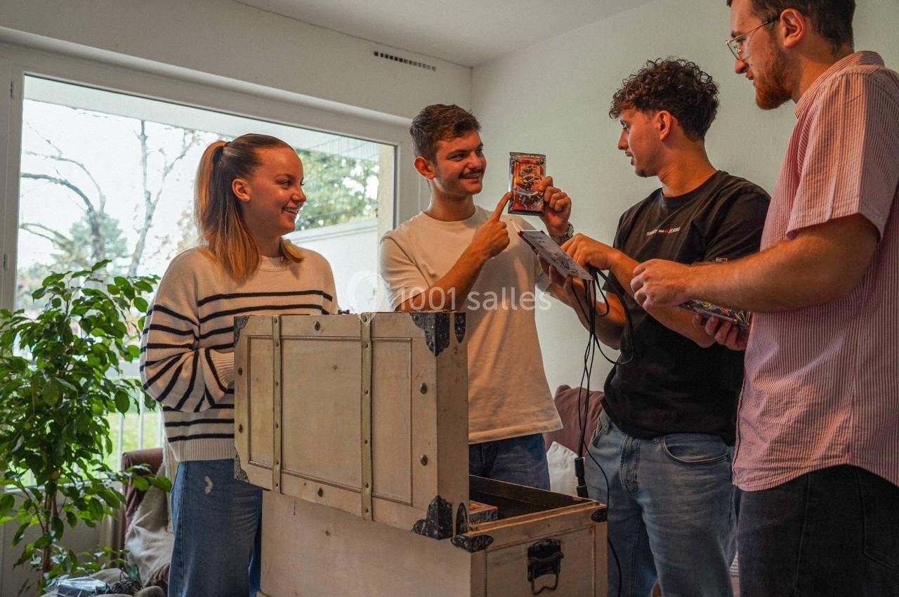 Quatre personnes discutent autour d'un coffre ouvert dans un salon lumineux, examinant des objets qu'il contient.