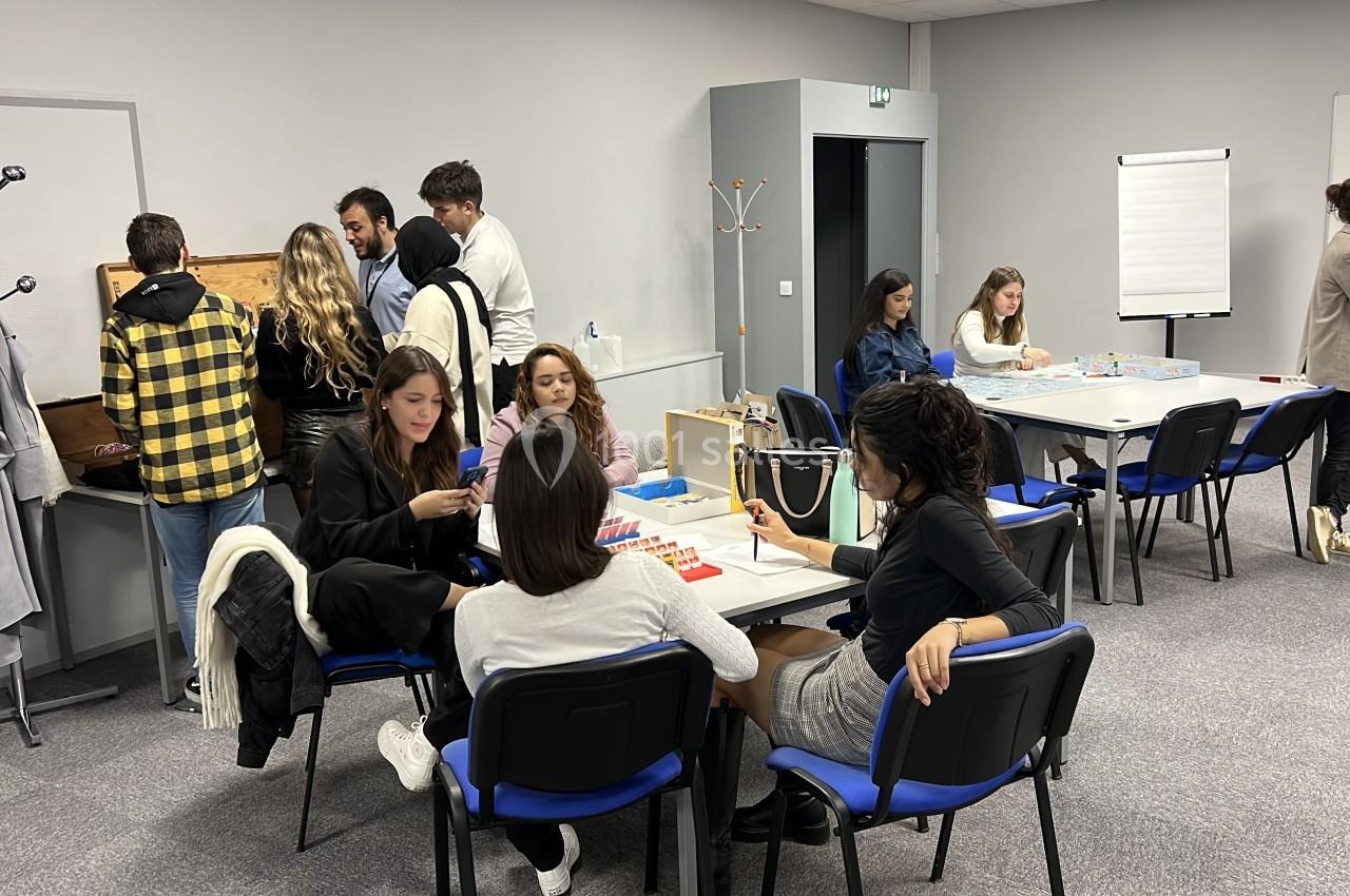 Des personnes participent à des activités en groupe dans une salle équipée de tables, chaises et tableau blanc.