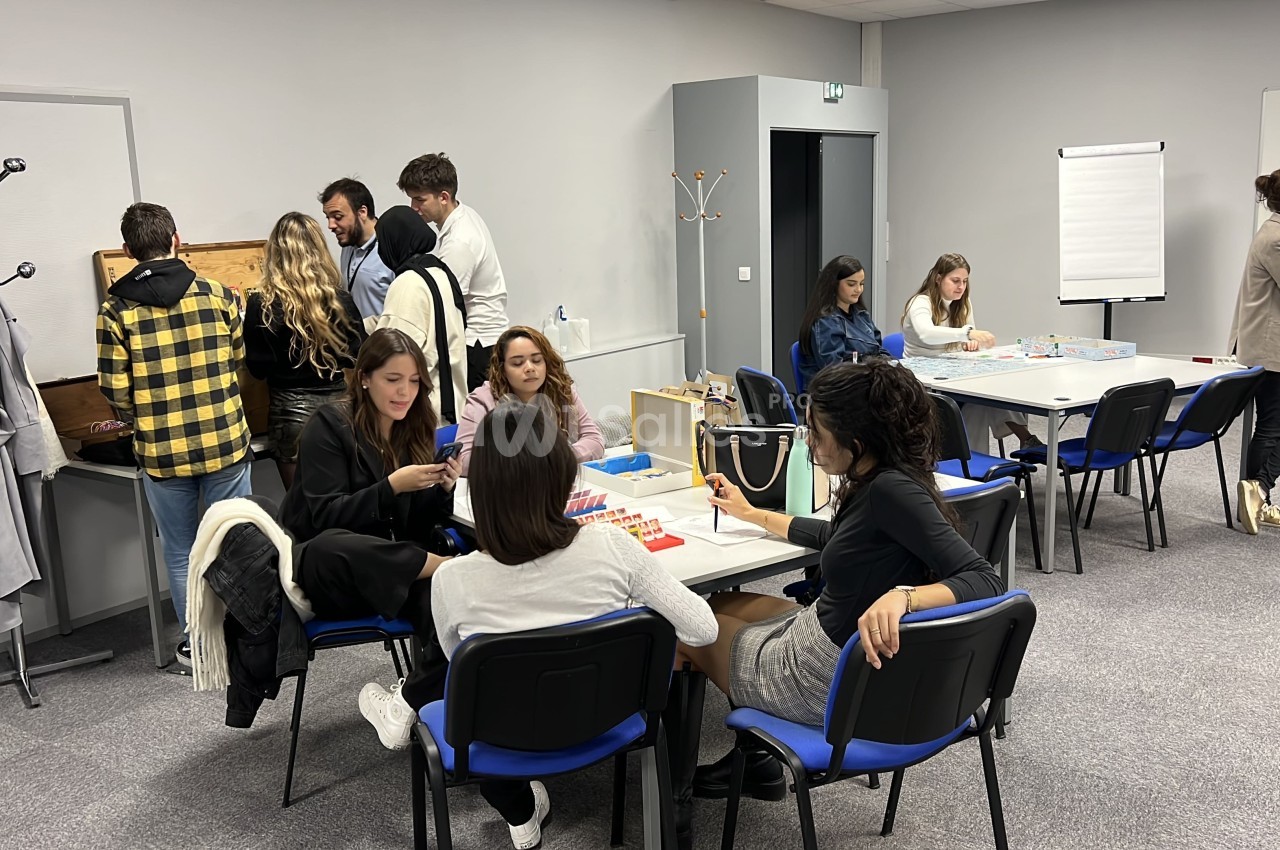 Des personnes participent à des activités en groupe dans une salle équipée de tables, chaises et tableau blanc.