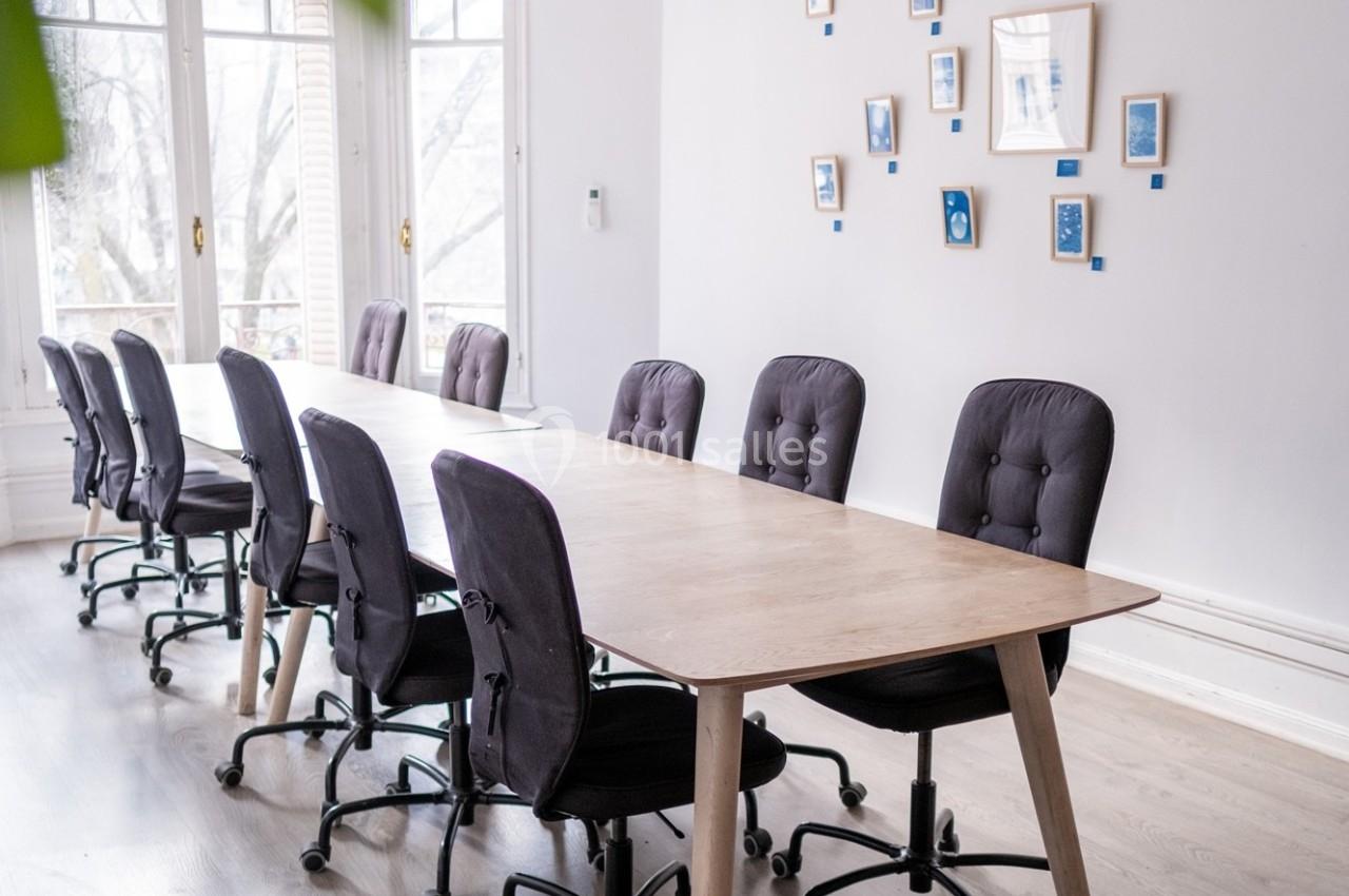 Salle de réunion lumineuse avec une grande table en bois, des chaises noires et des cadres accrochés au mur blanc.