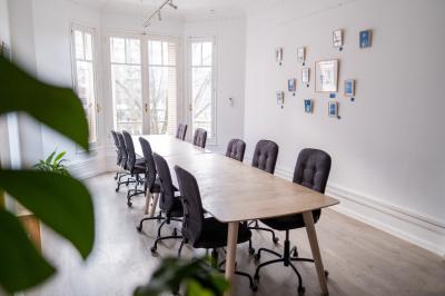 Salle lumineuse avec table ronde, chaises, tableau blanc, fauteuils et fenêtre donnant sur un extérieur arboré.