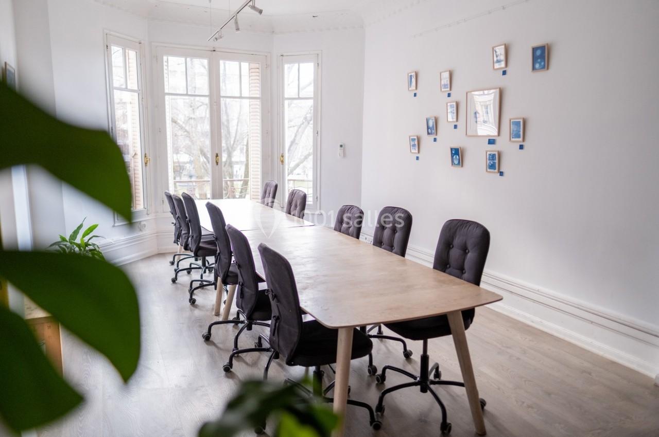 Salle de réunion lumineuse avec une grande table en bois, des chaises noires et des cadres accrochés au mur.
