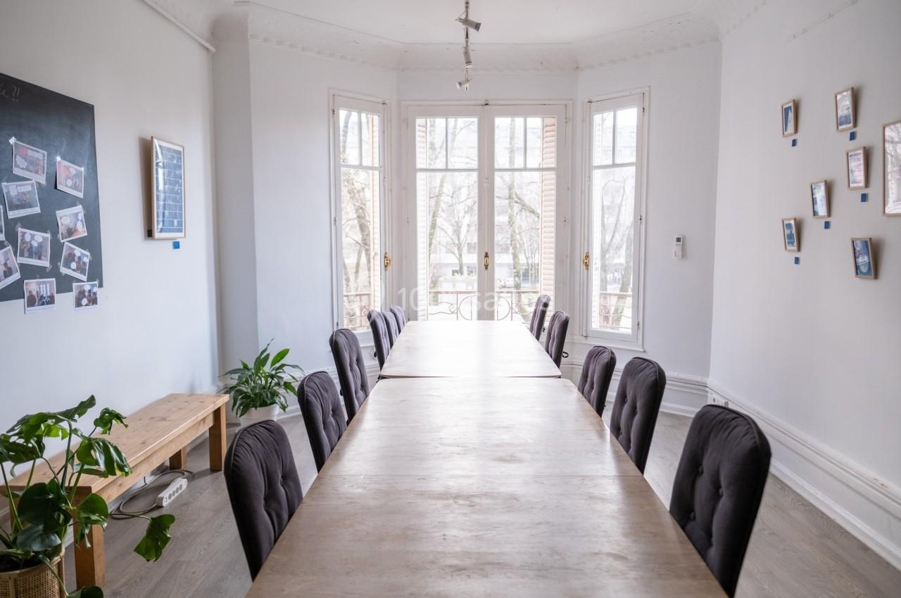 Salle lumineuse avec une grande table en bois entourée de chaises noires, des plantes et des cadres accrochés aux murs.