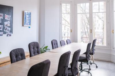 Salle lumineuse avec table ronde, chaises, tableau blanc, fauteuils et fenêtre donnant sur un extérieur arboré.