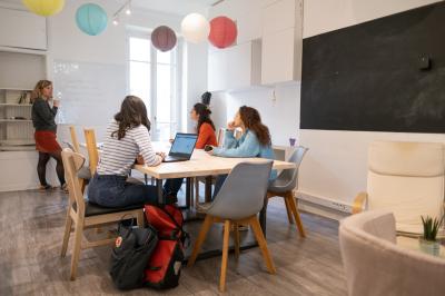Salle lumineuse avec table ronde, chaises, tableau blanc, fauteuils et fenêtre donnant sur un extérieur arboré.