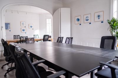 Salle lumineuse avec table ronde, chaises, tableau blanc, fauteuils et fenêtre donnant sur un extérieur arboré.