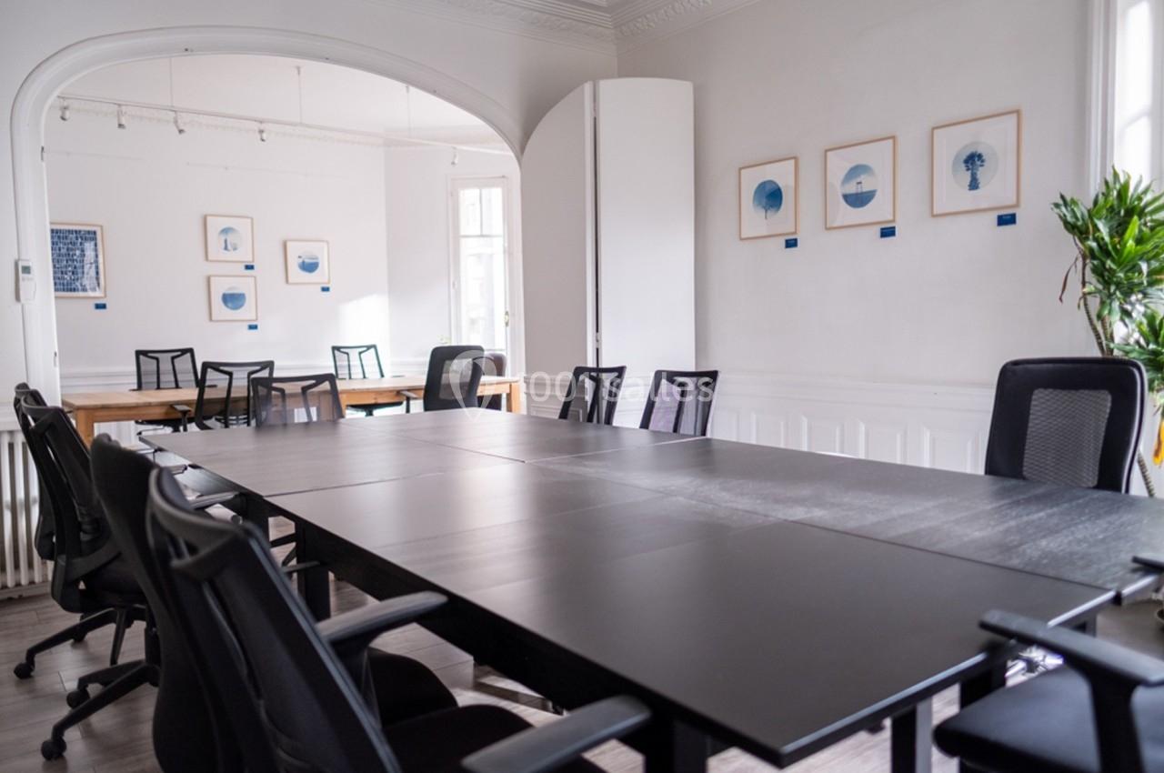 Salle de réunion lumineuse avec une grande table noire, des chaises de bureau et des cadres accrochés aux murs blancs.