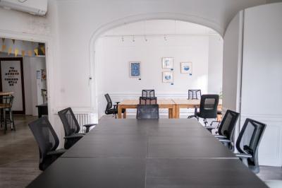 Salle lumineuse avec table ronde, chaises, tableau blanc, fauteuils et fenêtre donnant sur un extérieur arboré.