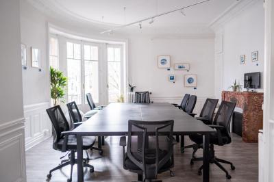 Salle lumineuse avec table ronde, chaises, tableau blanc, fauteuils et fenêtre donnant sur un extérieur arboré.