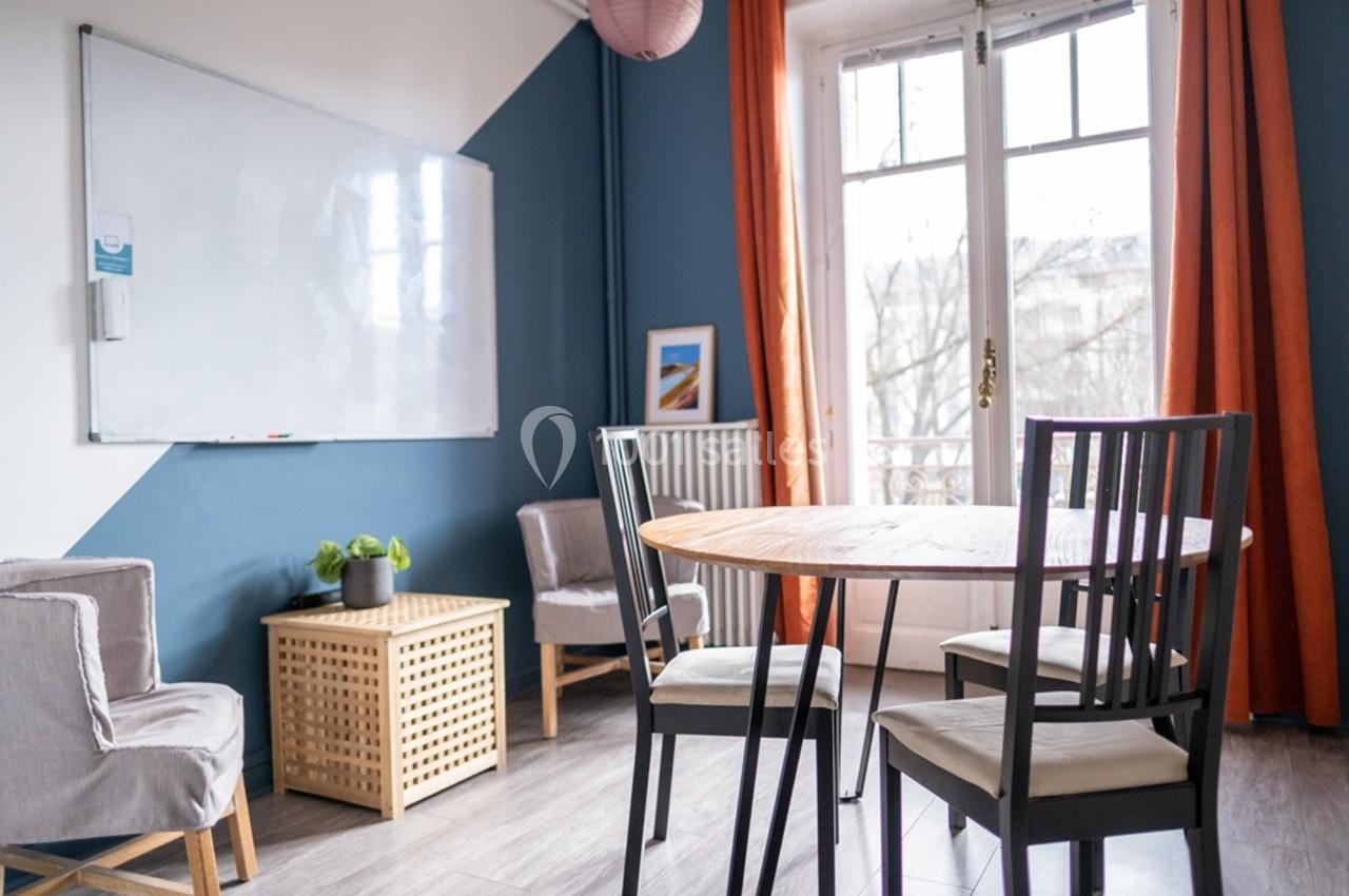 Salle lumineuse avec table ronde, chaises, tableau blanc, fauteuils et fenêtre donnant sur un extérieur arboré.