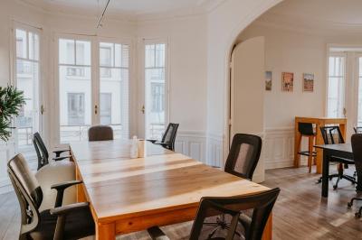 Salle lumineuse avec table ronde, chaises, tableau blanc, fauteuils et fenêtre donnant sur un extérieur arboré.