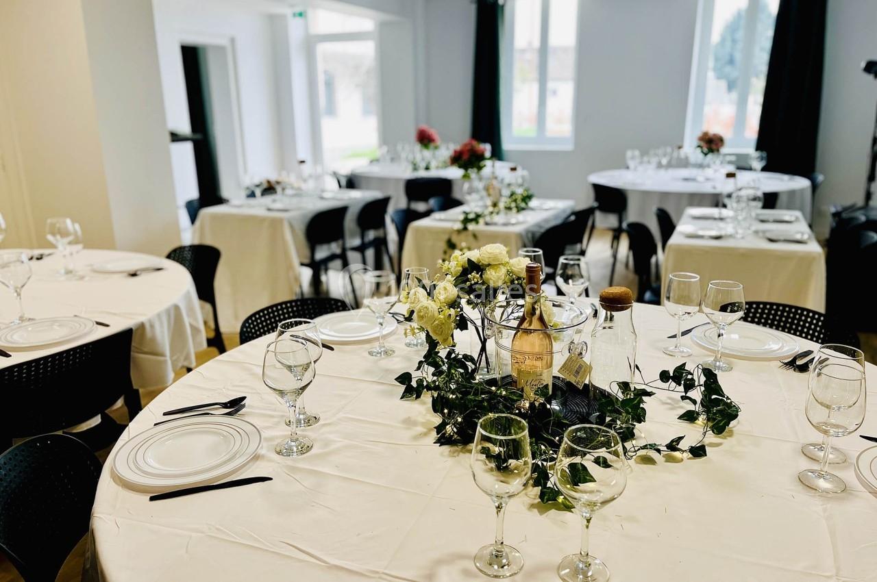 Tables rondes décorées avec nappes blanches, verres et centres de table fleuris dans une salle lumineuse.