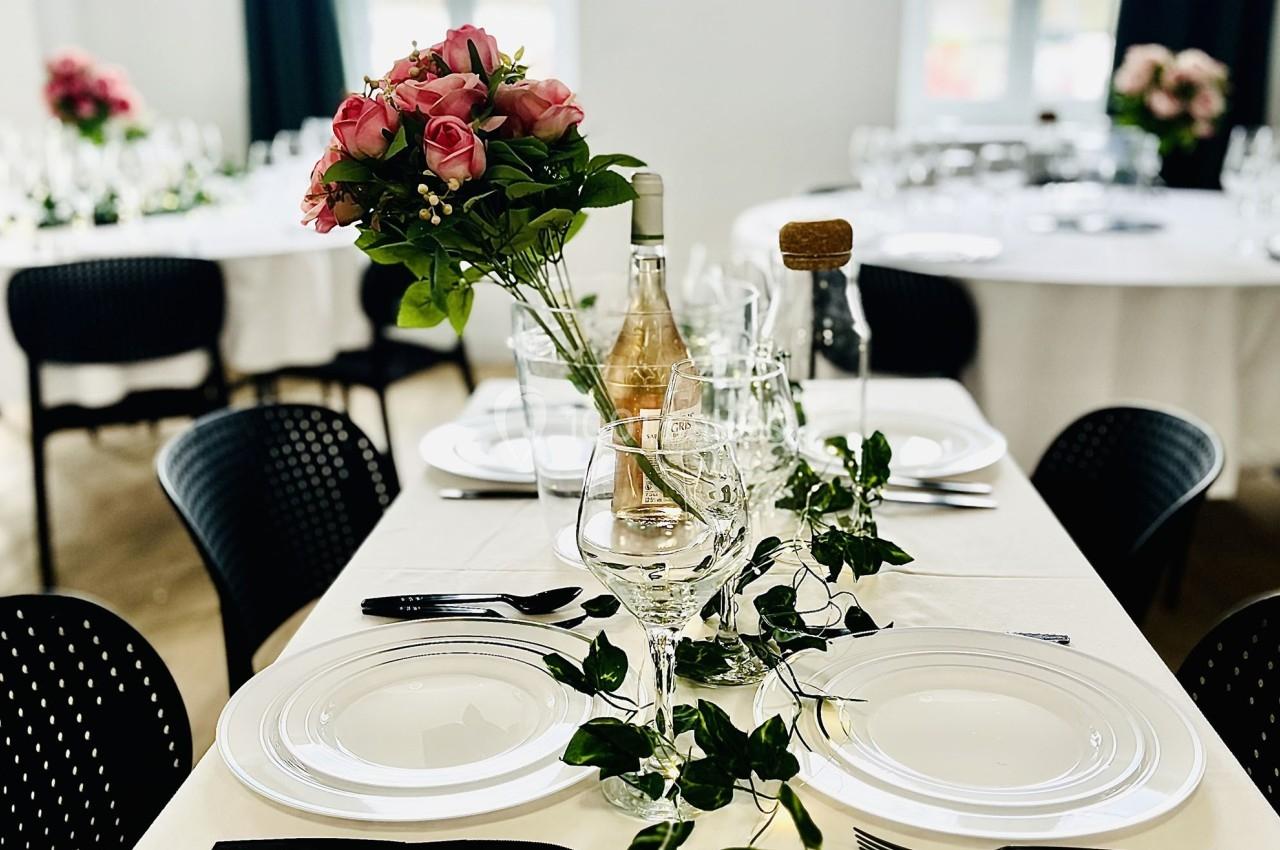 Table dressée avec nappes blanches, vaisselle élégante, décoration florale et guirlande végétale dans une salle lumineuse.