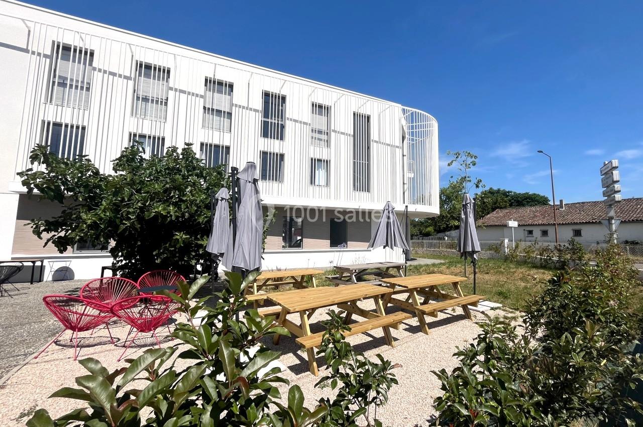 Espace extérieur avec tables en bois, parasols gris, chaises rouges et bâtiment moderne en arrière-plan.