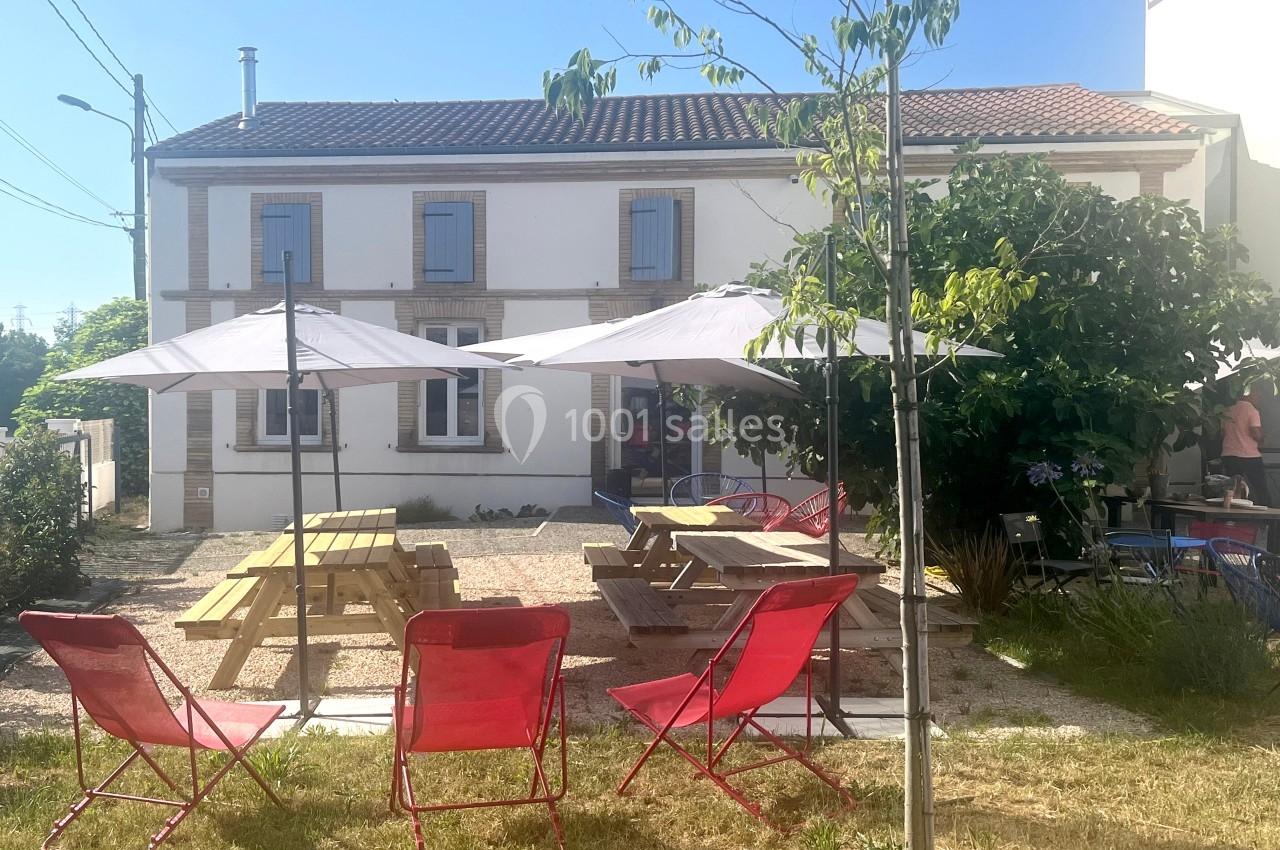 Façade d'une maison avec terrasse aménagée, tables en bois, parasols blancs et chaises rouges sur une pelouse.