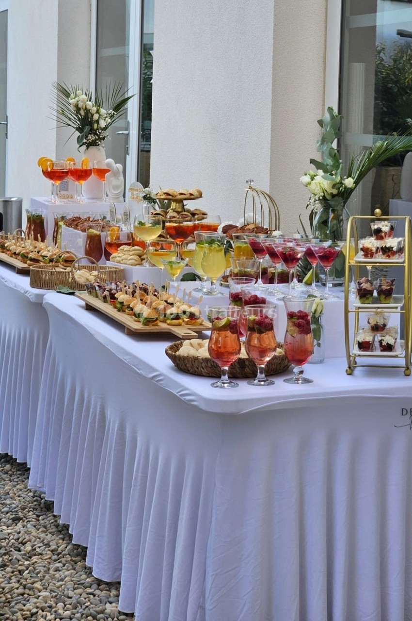 Buffet élégant en extérieur avec boissons, verrines, pâtisseries et décorations florales sur nappes blanches.