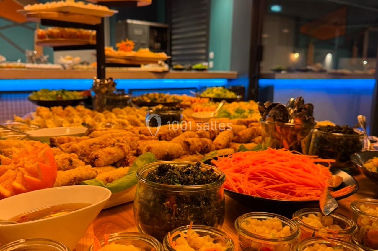 Buffet varié avec plats colorés, verrines, légumes et autres mets disposés sur une table en bois dans un cadre lumineux.