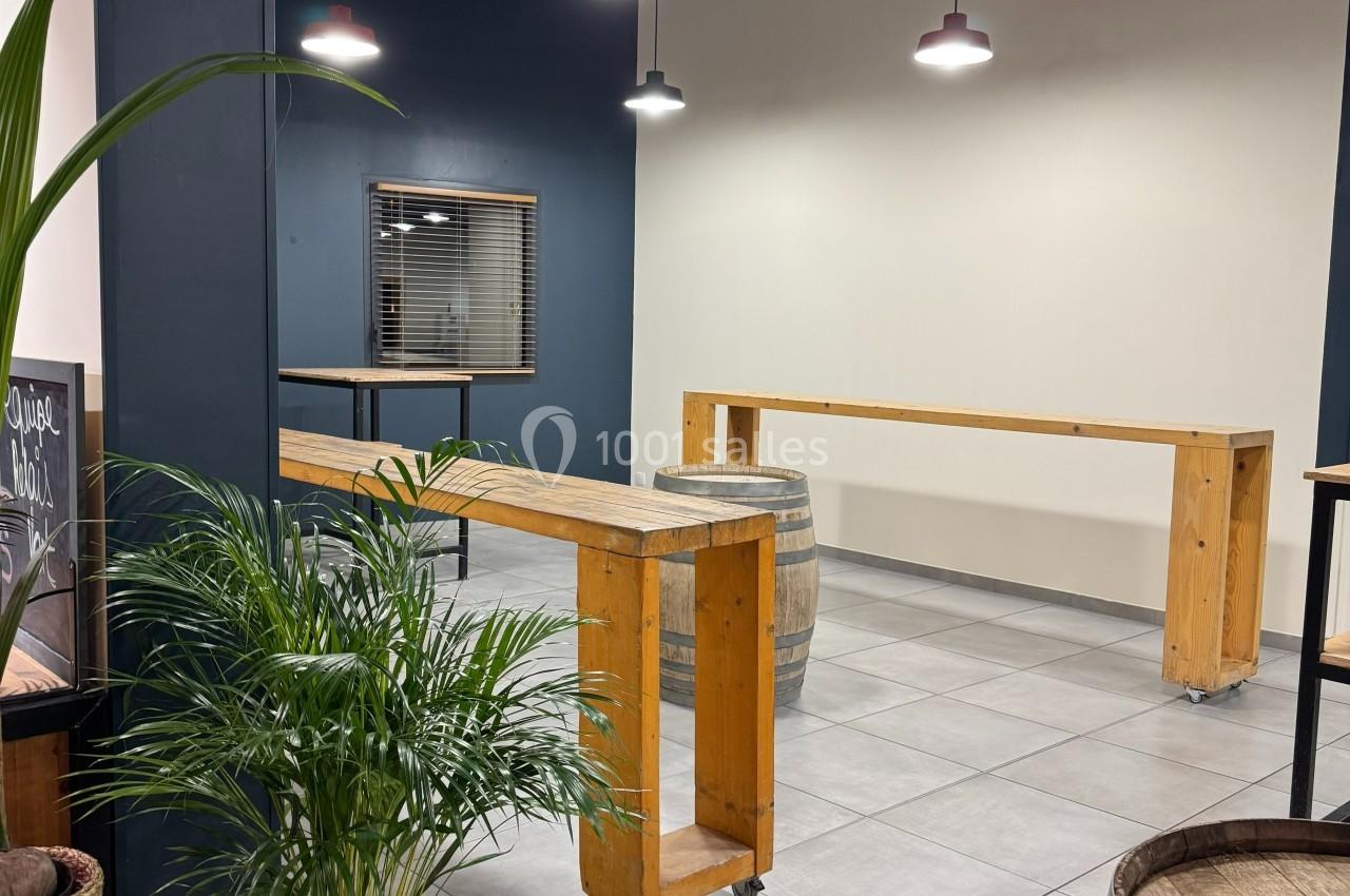 Salle moderne avec tables hautes en bois, tonneaux décoratifs, plantes vertes et éclairage suspendu.