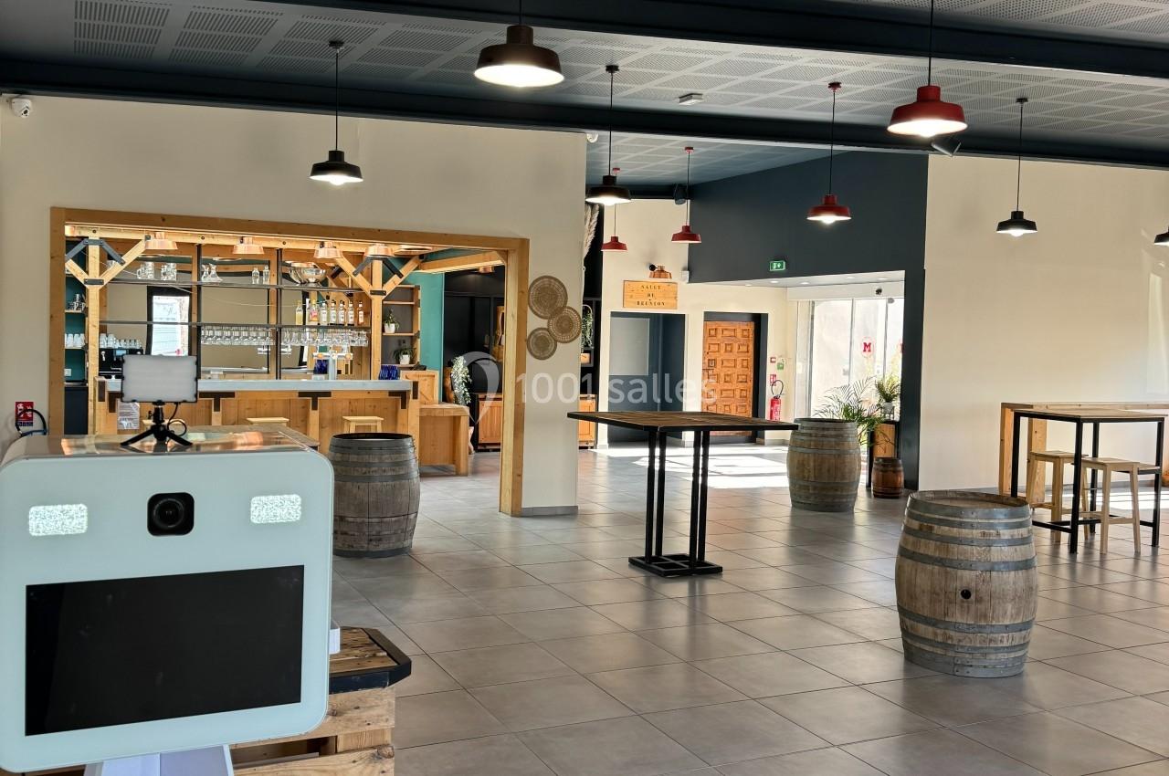 Salle lumineuse avec bar en bois, tables hautes, tonneaux décoratifs et un photobooth au premier plan.