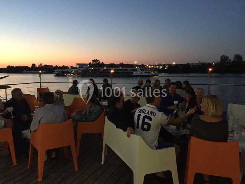 Groupe de personnes assises en terrasse au bord d'un fleuve au crépuscule, avec un bateau en arrière-plan.