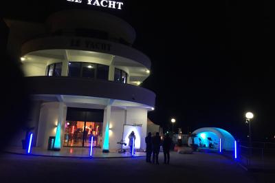 Façade éclairée d'un bâtiment nommé ’Le Yacht’ de nuit, avec des lumières bleues et un espace extérieur aménagé.