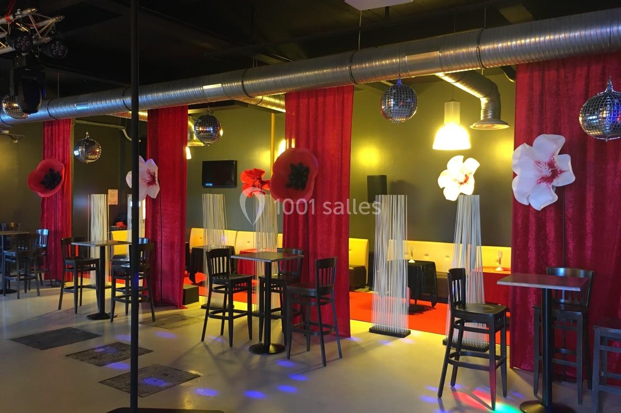 Salle décorée avec des tables hautes, rideaux rouges, fleurs murales et boules à facettes suspendues.