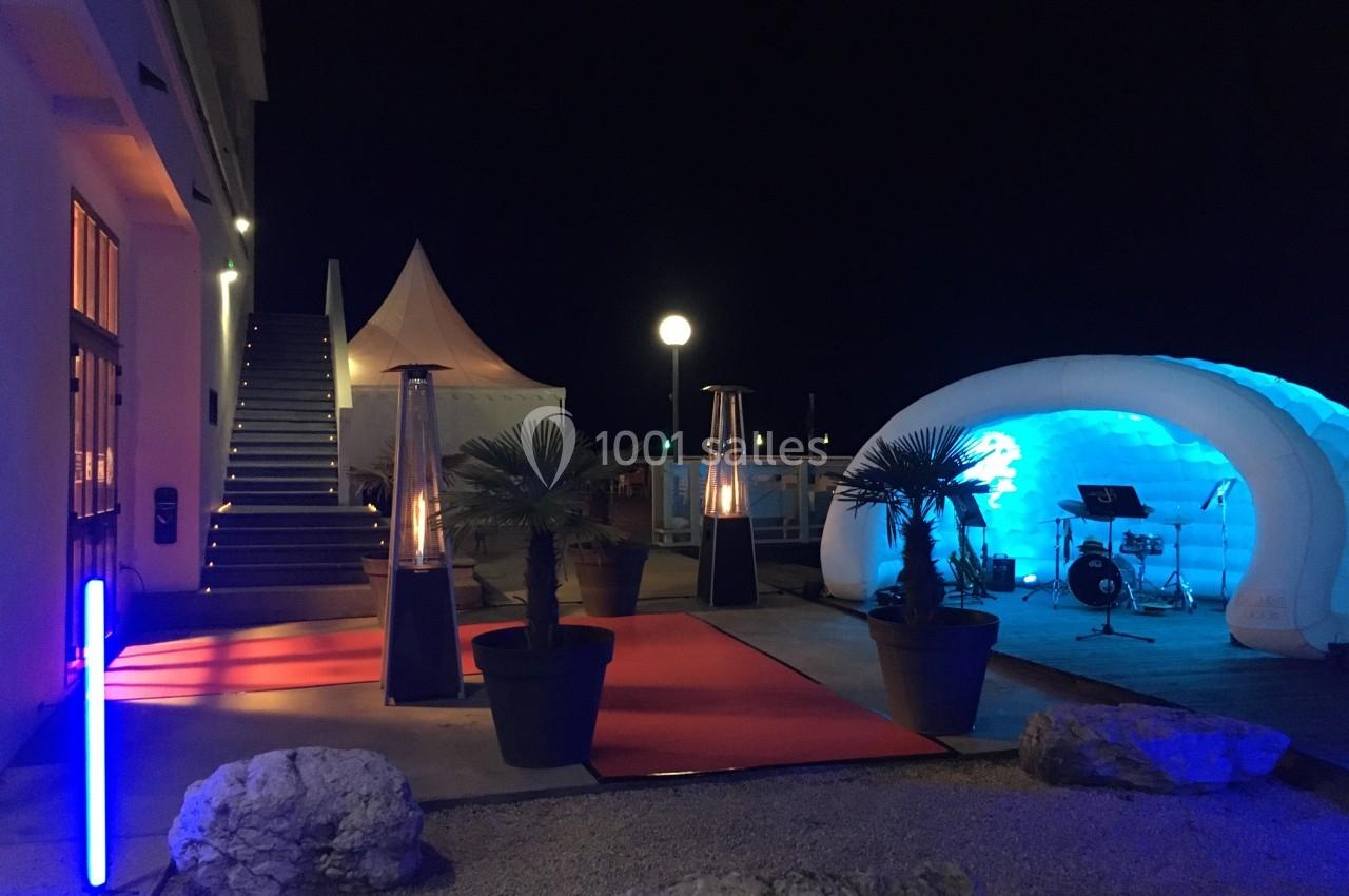 Entrée extérieure éclairée de nuit avec tapis rouge, palmiers en pot, chauffages d'appoint et tente gonflable blanche.