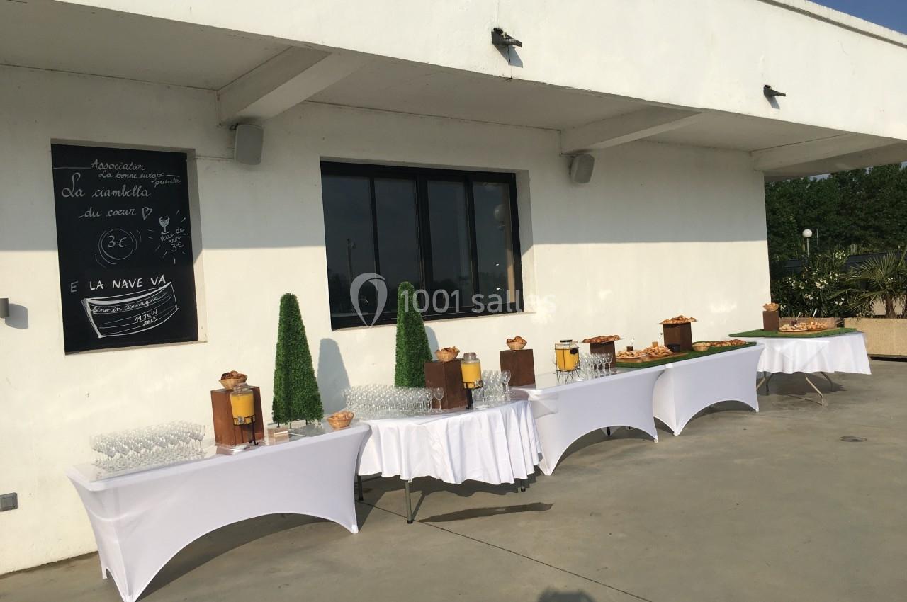 Buffet en extérieur avec tables dressées, boissons, verres et snacks disposés sous un mur blanc avec une ardoise décorative.