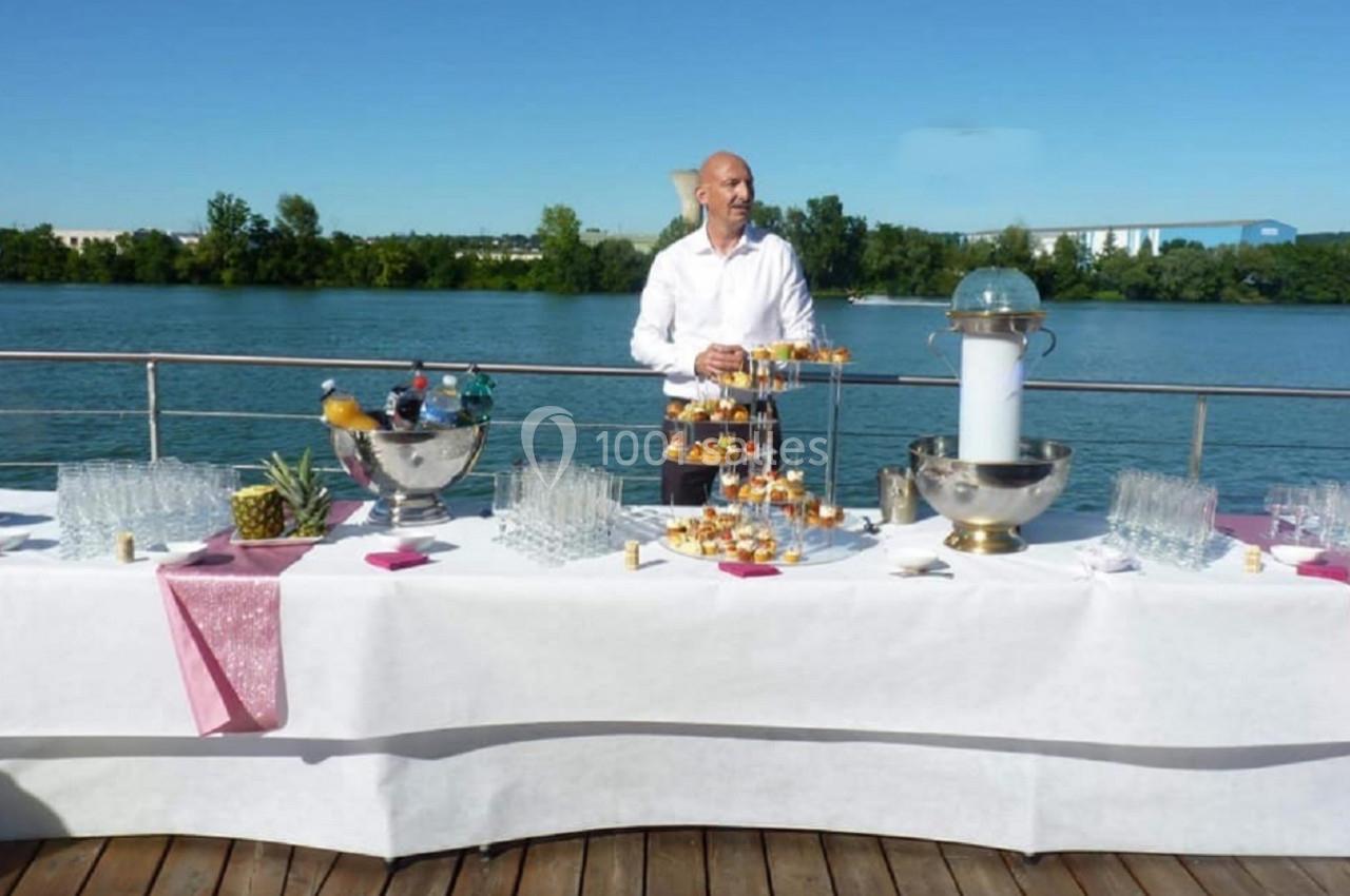Un homme debout derrière une table dressée avec des boissons et des amuse-bouches, sur un ponton au bord d'une rivière.