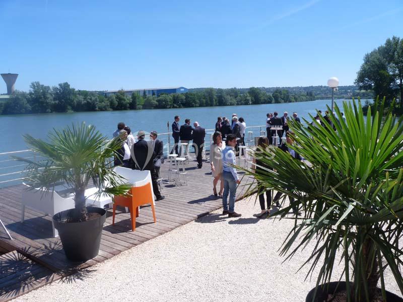 Personnes discutant sur une terrasse en bois au bord d'une rivière, entourée de plantes et sous un ciel dégagé.