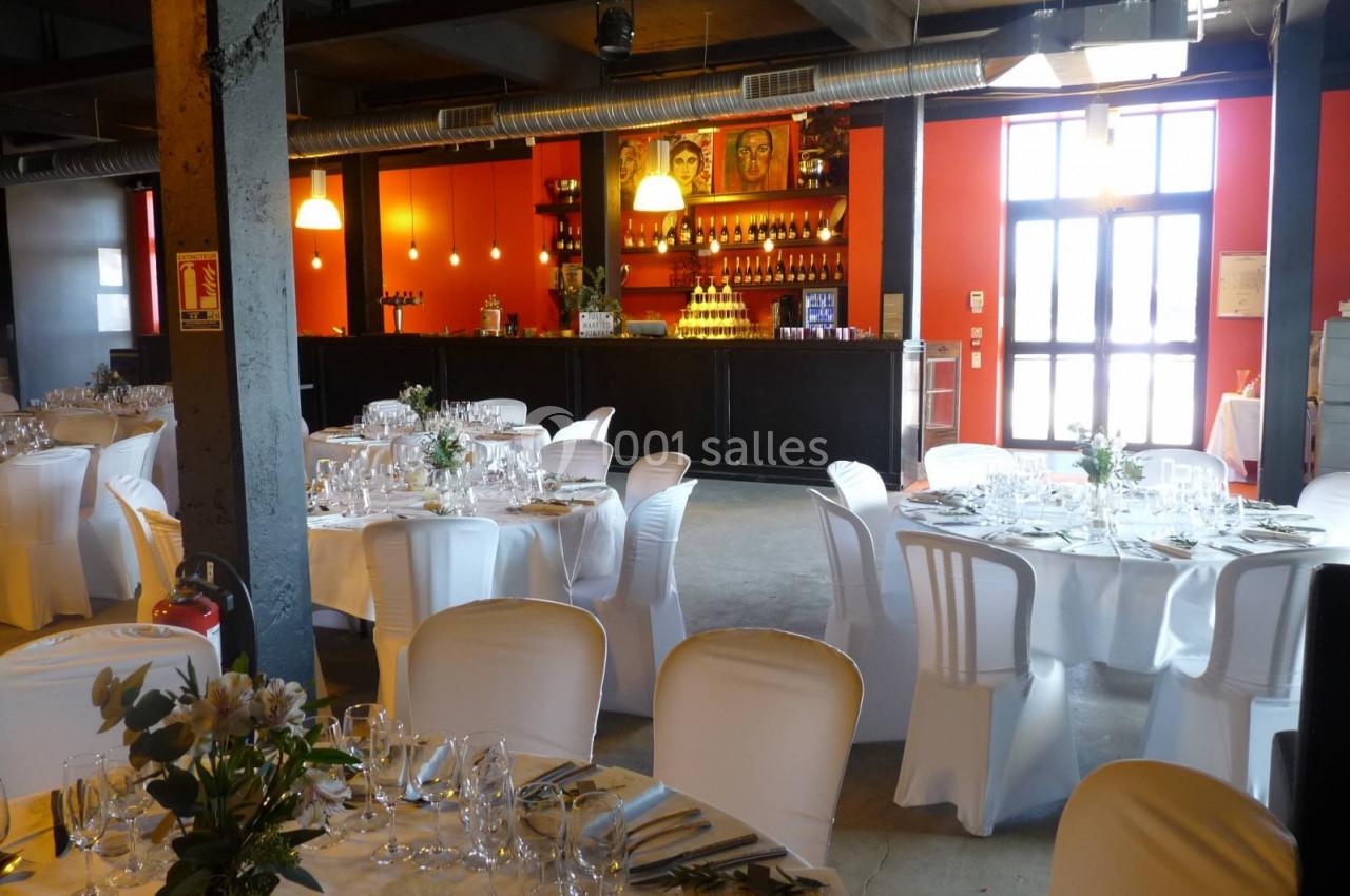 Salle de réception décorée avec des tables rondes dressées, chaises blanches et un bar en arrière-plan.