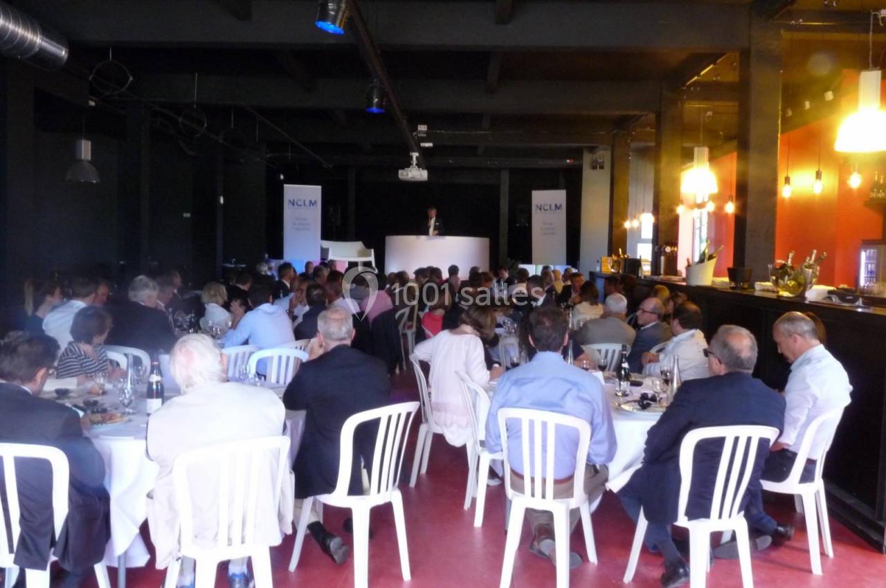 Salle de conférence avec des participants assis à des tables rondes, écoutant un intervenant sur une estrade.
