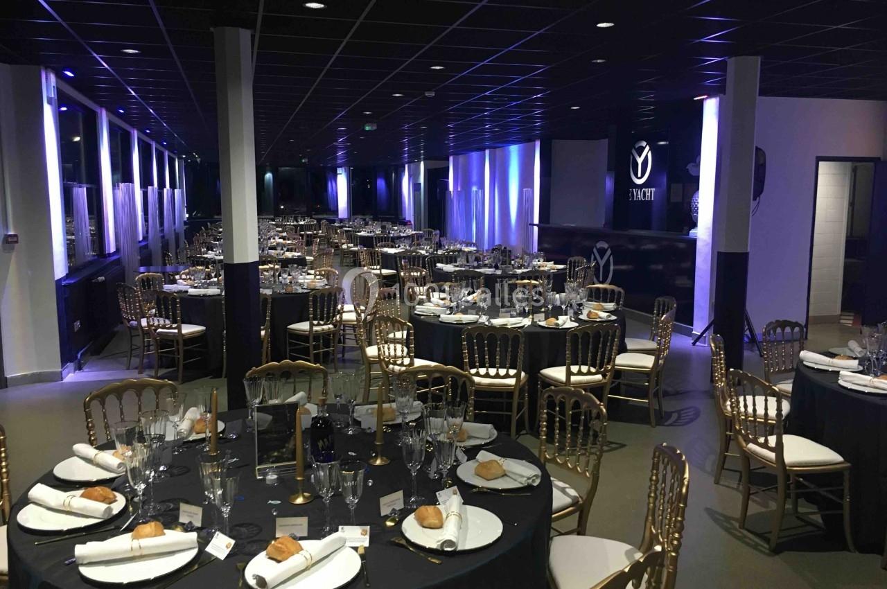 Salle de réception élégamment décorée avec tables rondes, nappes noires, chaises dorées et couverts dressés.