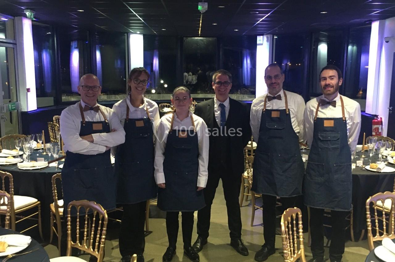 Équipe de serveurs en tabliers et homme en costume posant dans une salle de réception élégamment dressée.