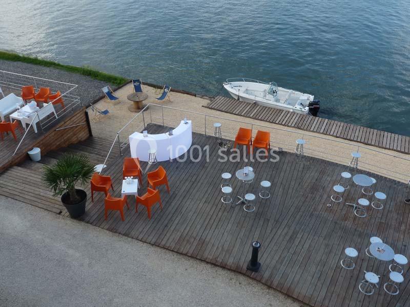 Terrasse en bois avec tables et chaises orange et blanches, bordée par un quai au bord de l'eau avec un bateau amarré.